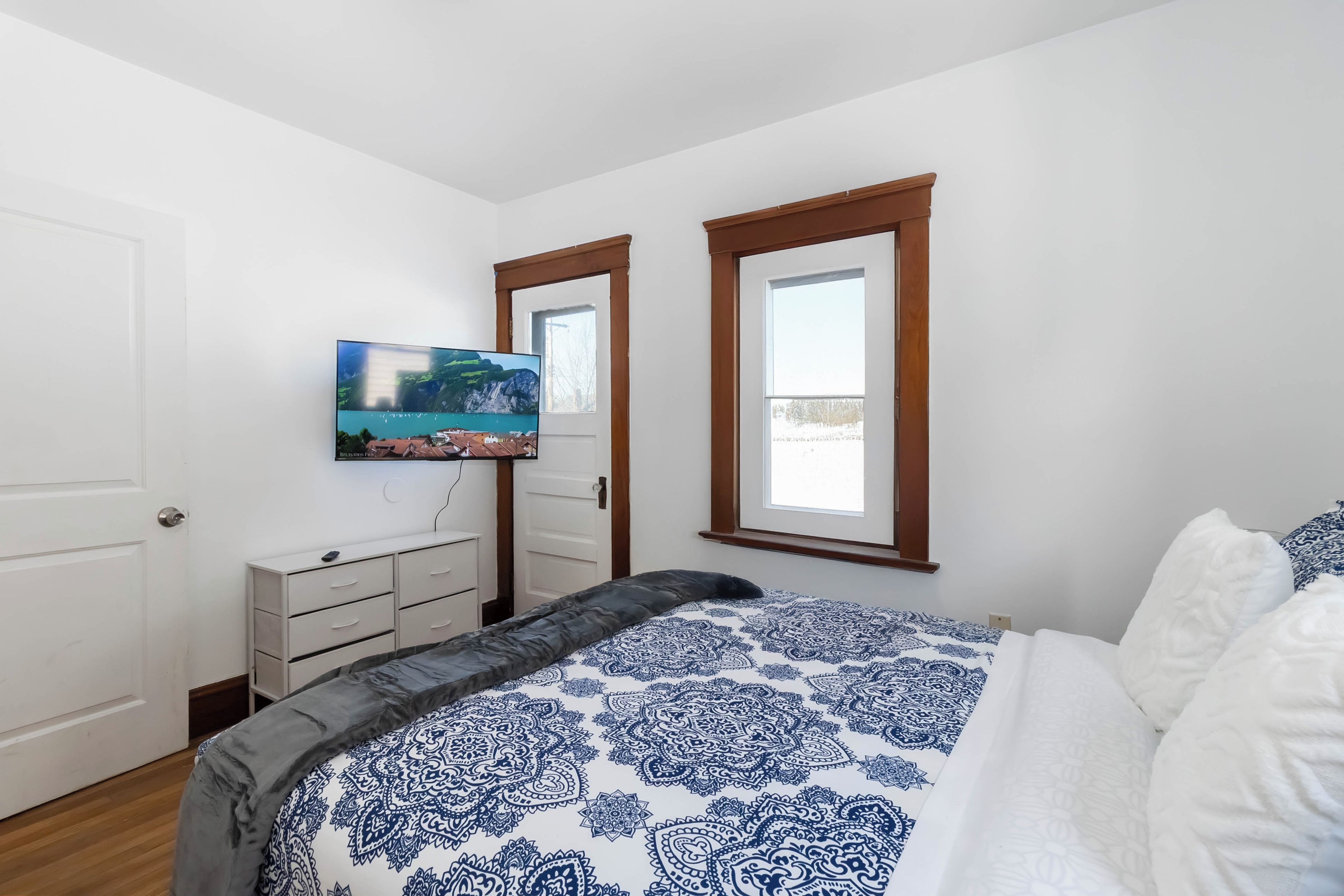A bedroom with a bed, a dresser, a television mounted on the wall, and two windows.