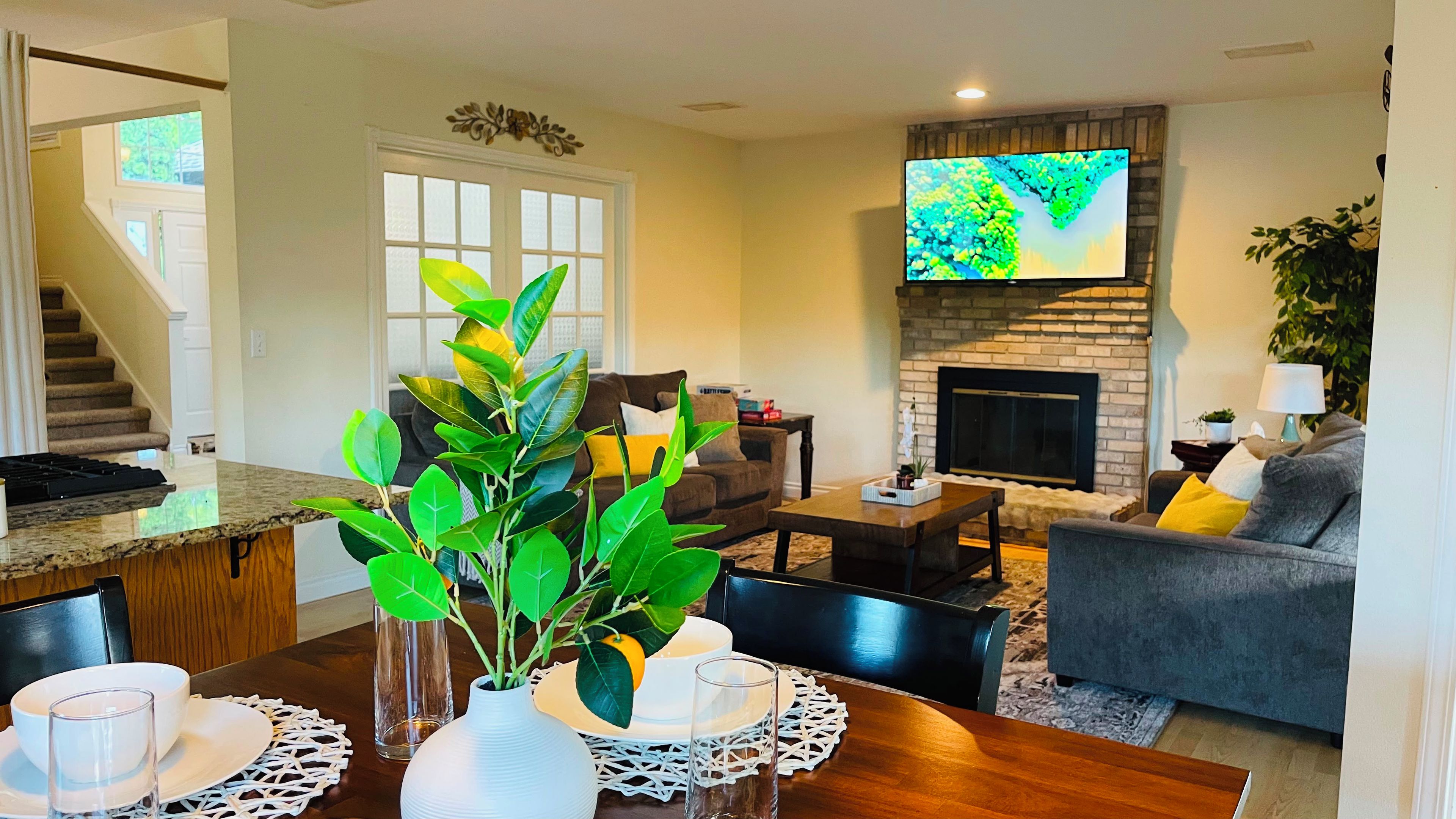Una sala de estar con chimenea, un televisor de pantalla plana, un comedor con una mesa lista para dos personas, y una escalera que conduce a otro nivel.
