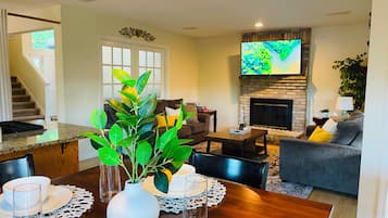 Una sala de estar con chimenea, un televisor de pantalla plana, un comedor con una mesa lista para dos personas, y una escalera que conduce a otro nivel.