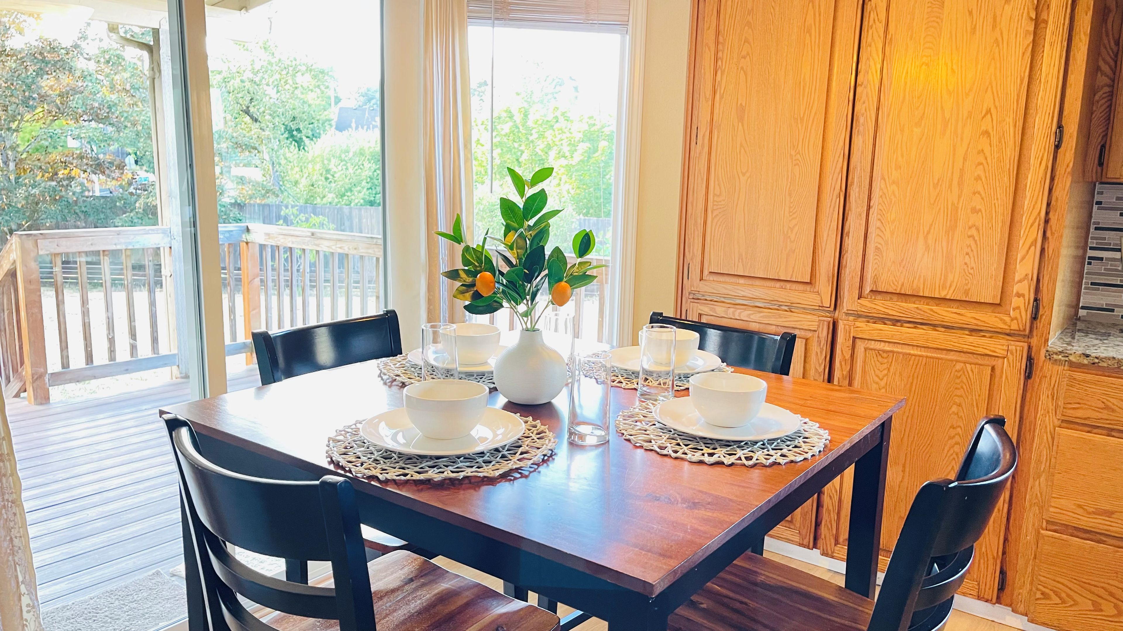 Une table à manger dressée avec de l’assiette blanche, des bols et un vase contenant des feuilles vertes et des fruits oranges.