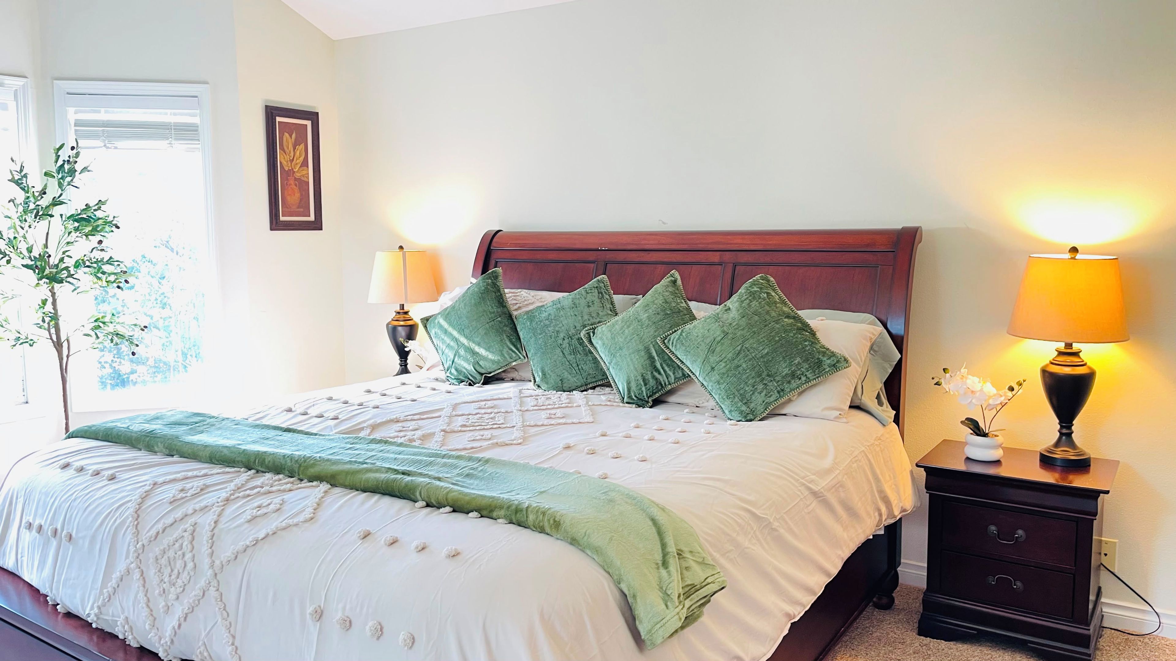 A neatly made bed with a green throw blanket and pillows, bedside lamps, a nightstand, and a potted plant.