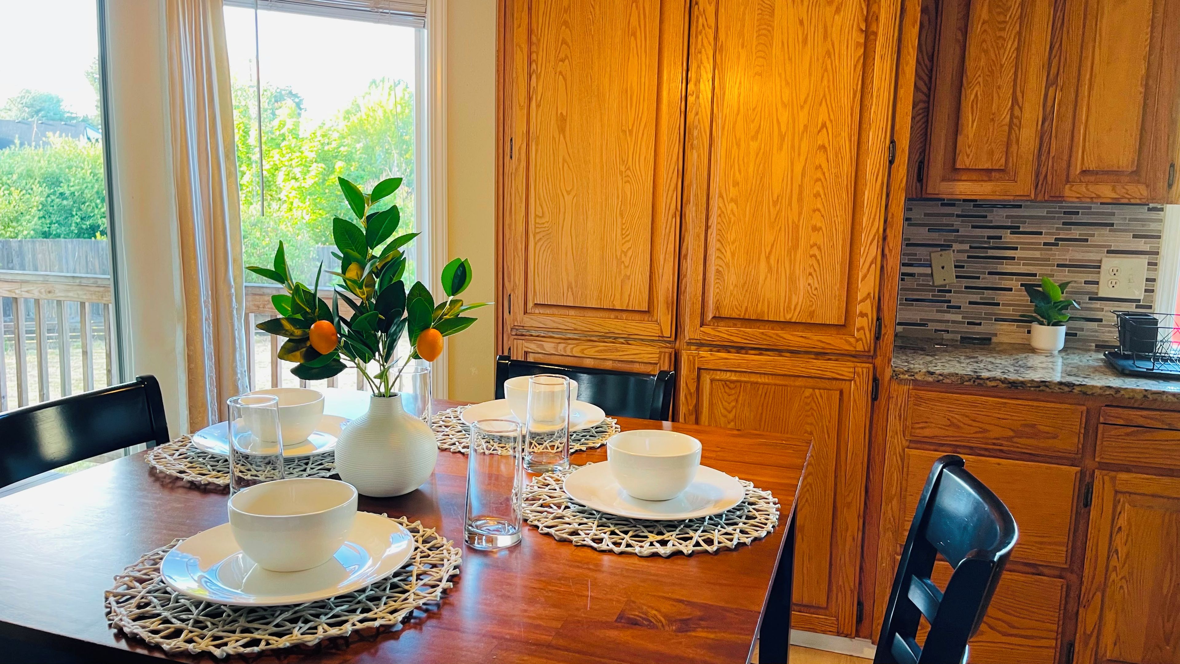 A dining area with a wooden table set for four, surrounded by wooden cabinets and a kitchen area with a tiled backsplash.