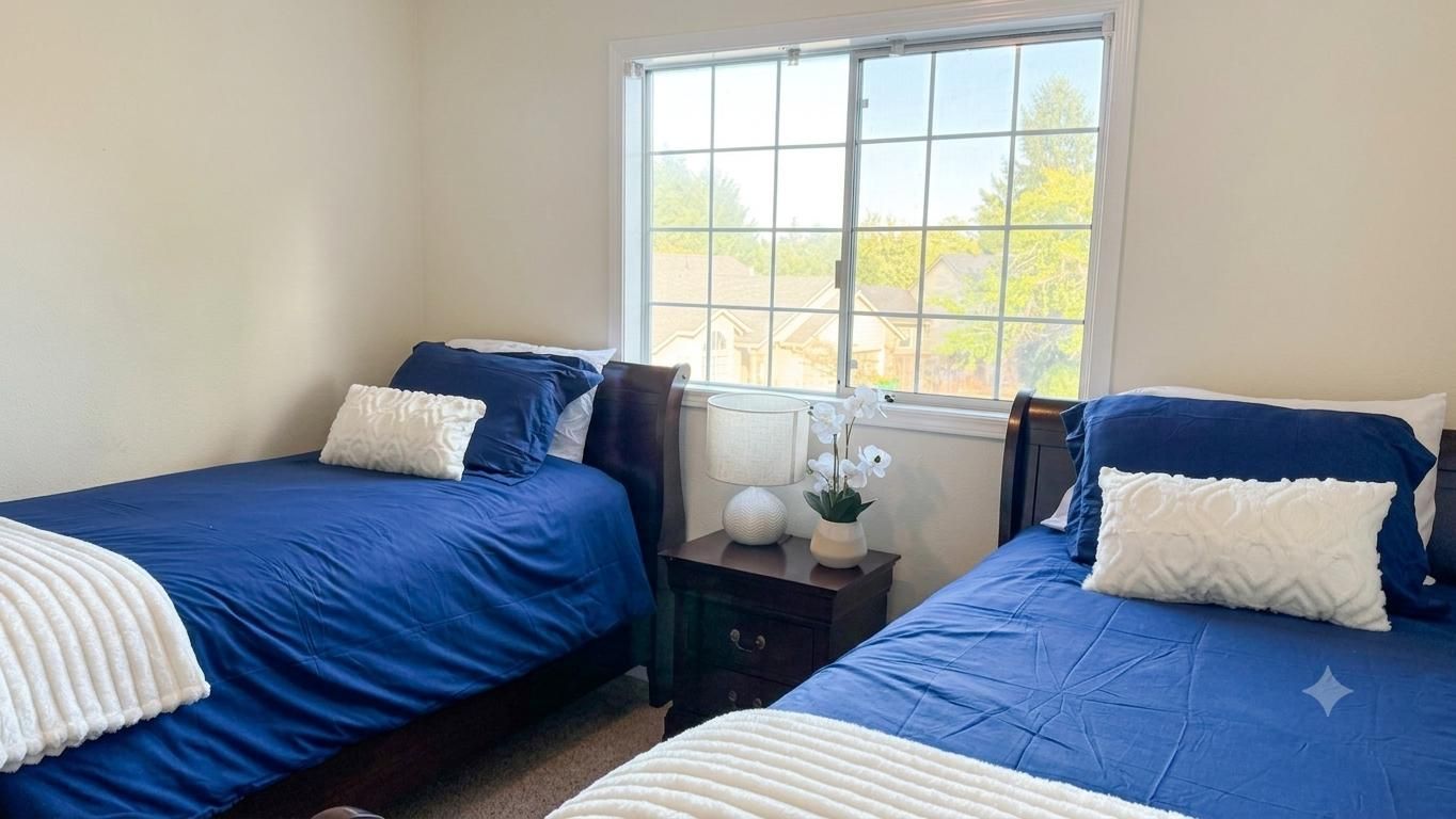 Une chambre avec deux lits, une table de chevet, une lampe et une fenêtre donnant sur des maisons.