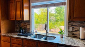 A kitchen with wooden cabinets, a granite countertop, a double sink, and a window with a view of trees.