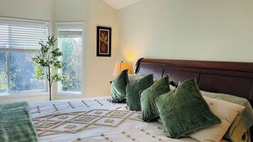 A bedroom with a bed, pillows, a lamp, a window with blinds, and a plant.