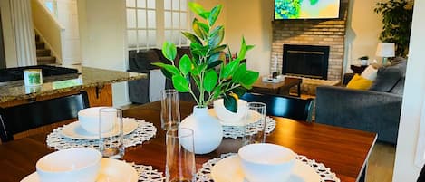 Un coin repas avec une table en bois, dressée pour quatre personnes et entourée de chaises, et un téléviseur fixé au-dessus d’une cheminée.