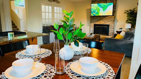 A dining area with a wooden table set for four, surrounded by chairs, with a television mounted above a fireplace.