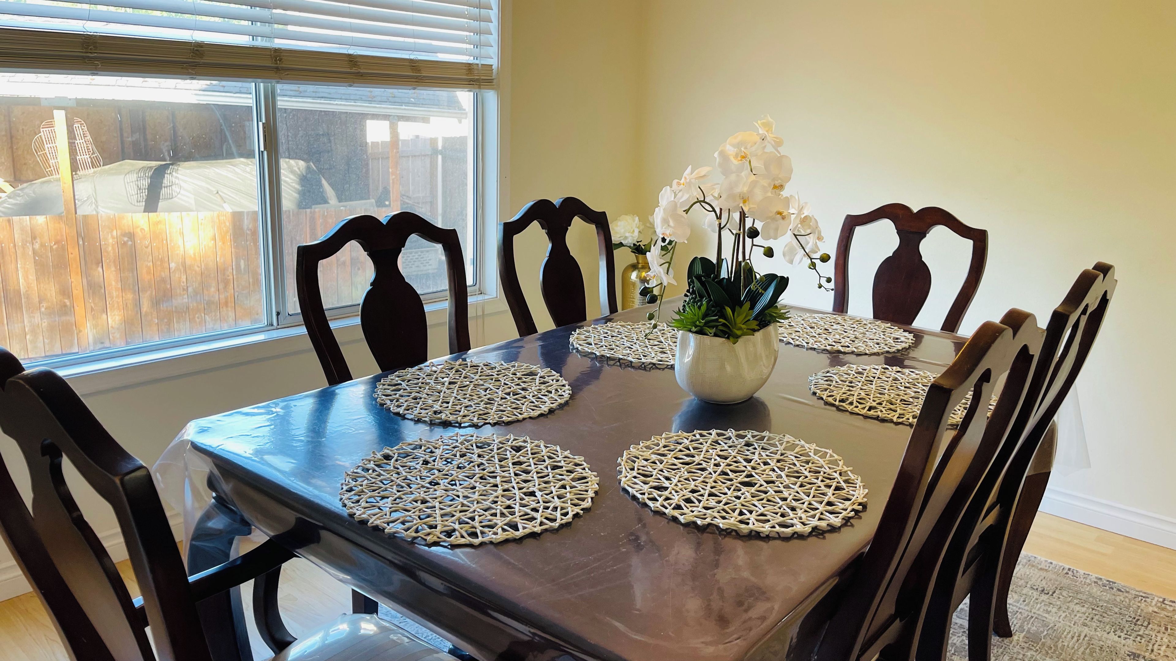 Une table à manger avec des sets de table en fibres tressées, des chaises et une plante en pot ornée de fleurs blanches.