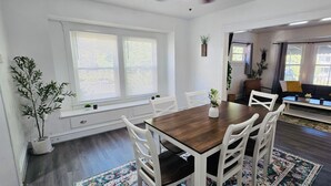 Ein Essbereich mit Holztisch und weißen Stühlen, einer Couch und einem Fenster mit Jalousien.