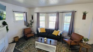 Ein Wohnzimmer mit einer Couch, zwei Sesseln, einem Couchtisch und einem Kamin. Es gibt Fenster mit Vorhängen, einen Fernseher und eine Pflanze an der Wand.