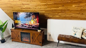 A living room with a fireplace, a television on a wooden stand, and a bench with decorative pillows. - Lakewood Cabin-style third floor Getaway 2p2 (Lakewood)