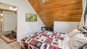 Ett sovrum med ett sluttande tak, en säng med ett mönstrat täcke, en tavla på väggen och en dörr som leder till ett annat rum.