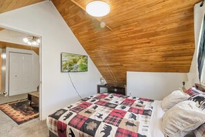 A bedroom with a sloped ceiling, a bed with a patterned quilt, a painting on the wall, and a door leading to another room. - Lakewood Cabin-style third floor Getaway 2p2 (Lakewood)