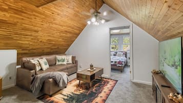 A loft bedroom with a sloped ceiling, a wooden ceiling, a ceiling fan, a bed with a forest-themed bedspread, a brown leather sofa with decorative pillows, and a television on a wooden stand.