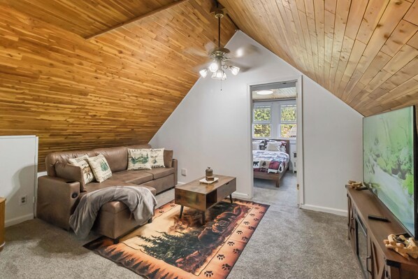 A loft bedroom with a sloped ceiling, a wooden ceiling, a ceiling fan, a bed with a forest-themed bedspread, a brown leather sofa with decorative pillows, and a television on a wooden stand.