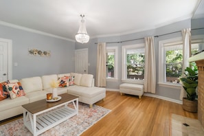 Una sala de estar con un sofá modular, una mesa de centro, una araña y ventanas amplias con cortinas.