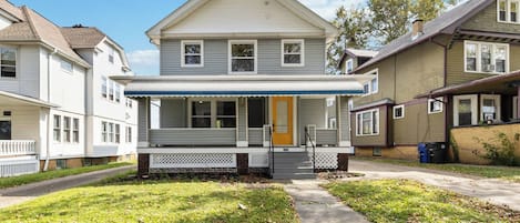 A two-story residential building with a front porch, a visible garage door, and a pathway leading to the entrance.