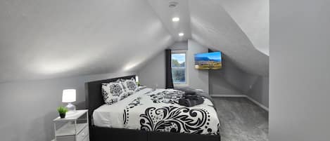 A bedroom with a bed, a nightstand, a lamp, a plant, and a TV mounted on the wall.