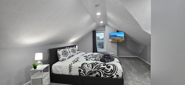 Ein Schlafzimmer mit einem Bett, einem Nachttisch, einer Lampe, einer Pflanze und einem an der Wand montierten Fernseher.