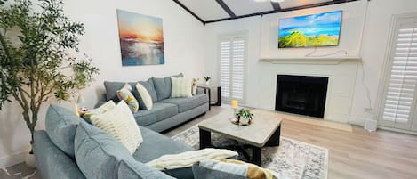 Een woonkamer met een grijze bank, een open haard, een televisie en een schilderij aan de muur.