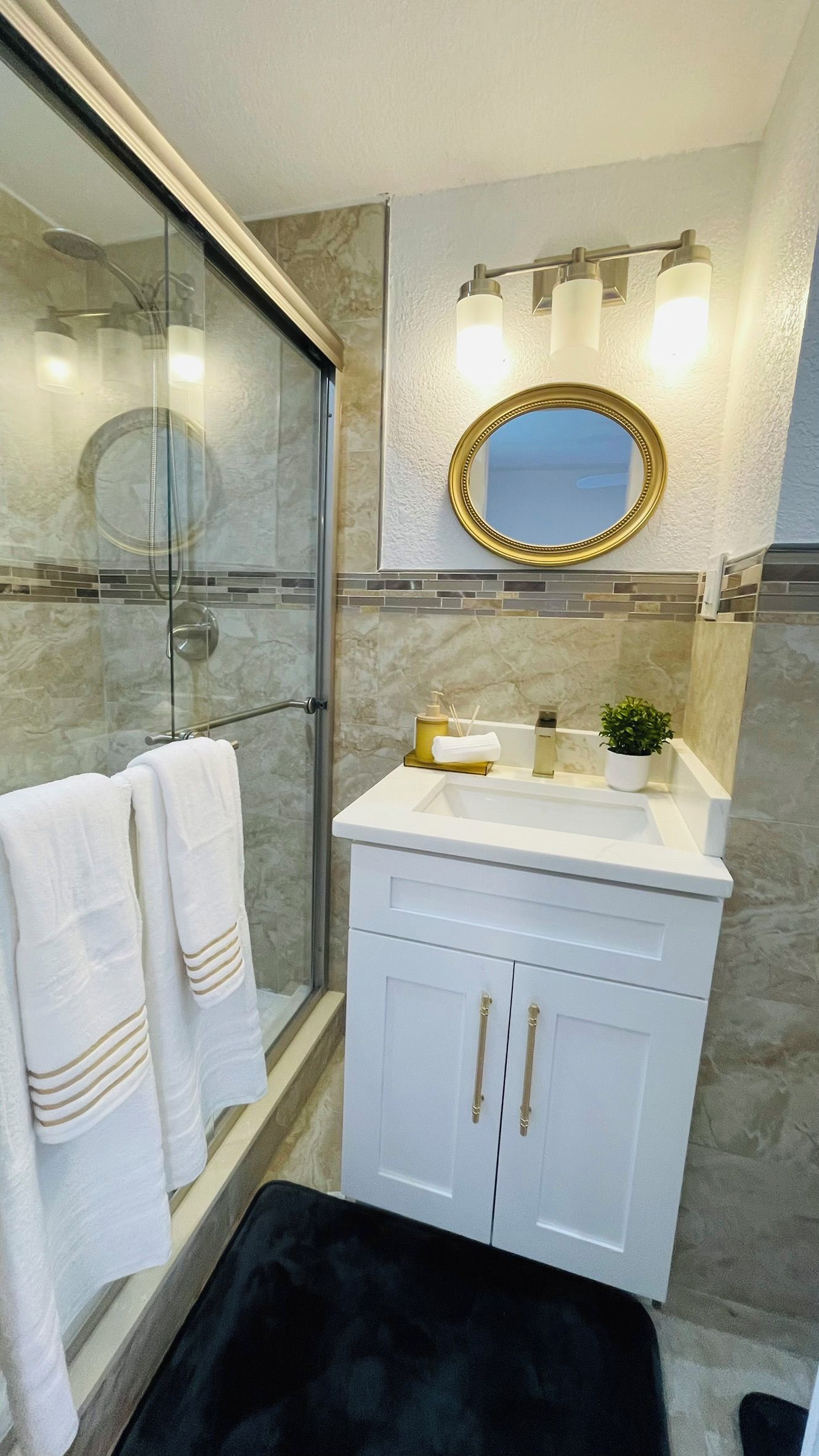 Et moderne badeværelse med et brusekabine i glas, et hvidt toiletbord med et spejl med guldramme og en sort badeværelsesmåtte.