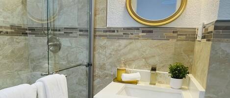 A modern bathroom with a glass shower enclosure, a white vanity with a gold-framed mirror, and a black bath mat.