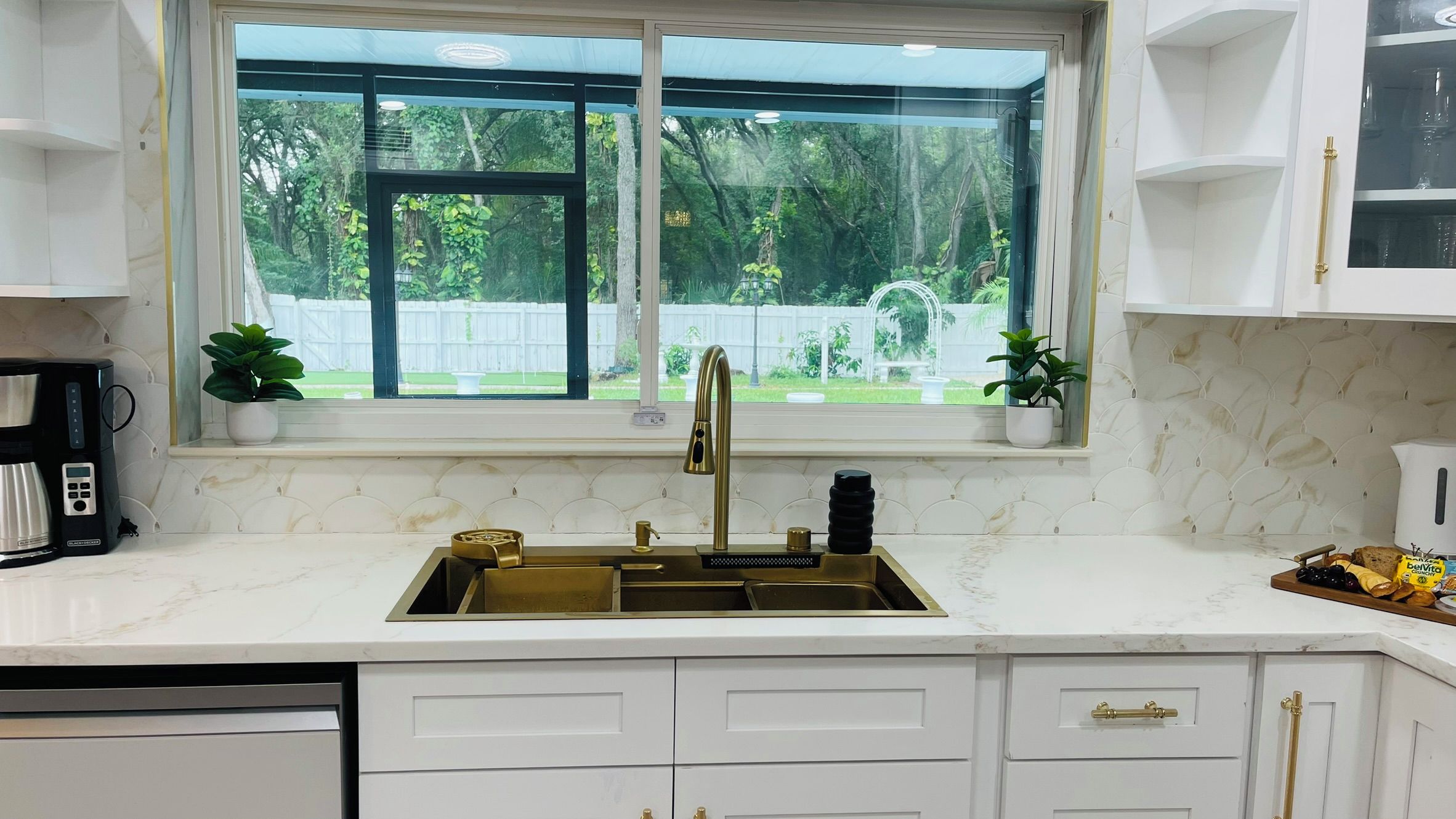 A kitchen with a large window, white cabinetry, a gold faucet, and a coffee maker.