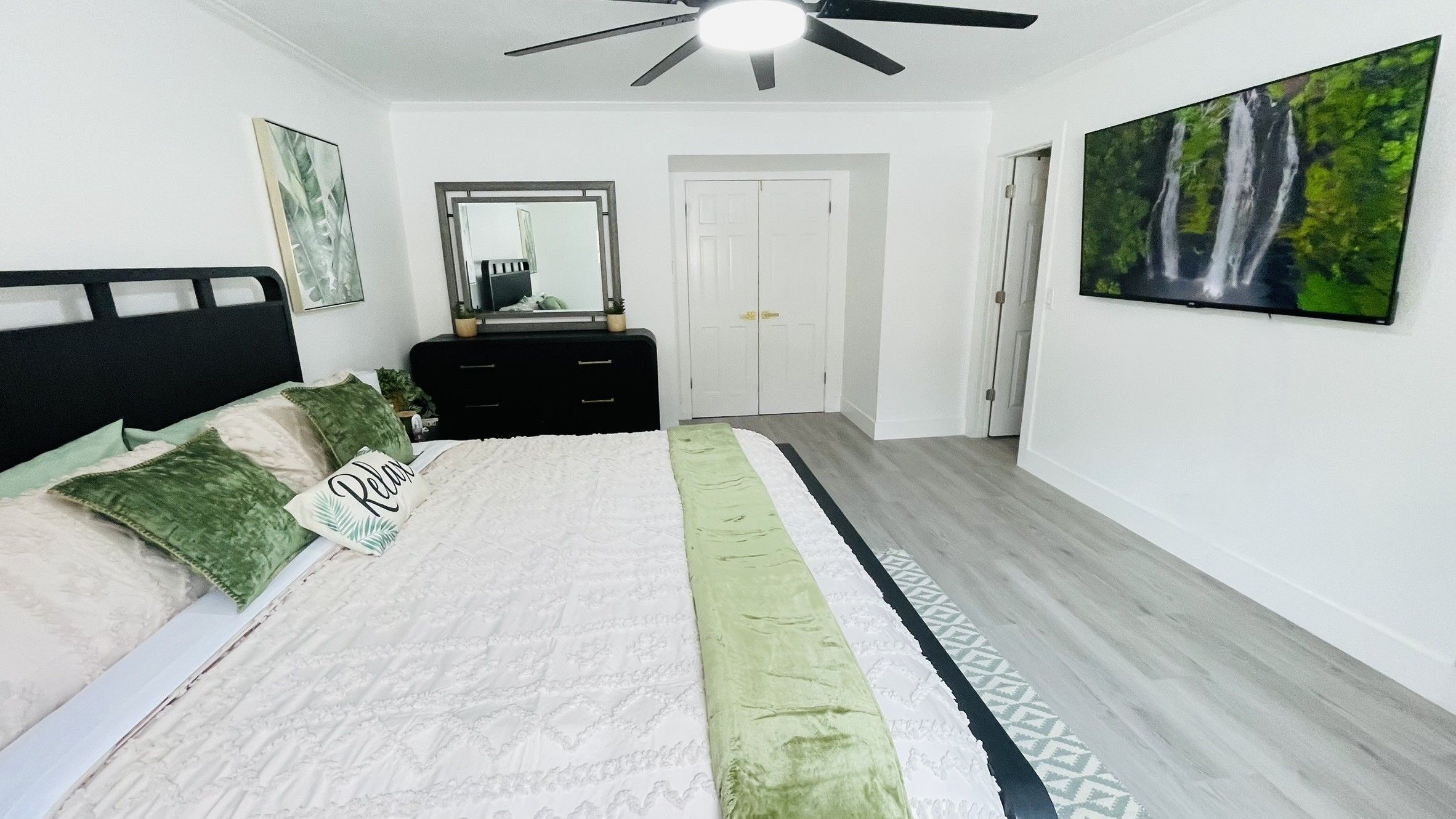 A bedroom with a bed, a dresser, a ceiling fan, a door, and a painting on the wall.