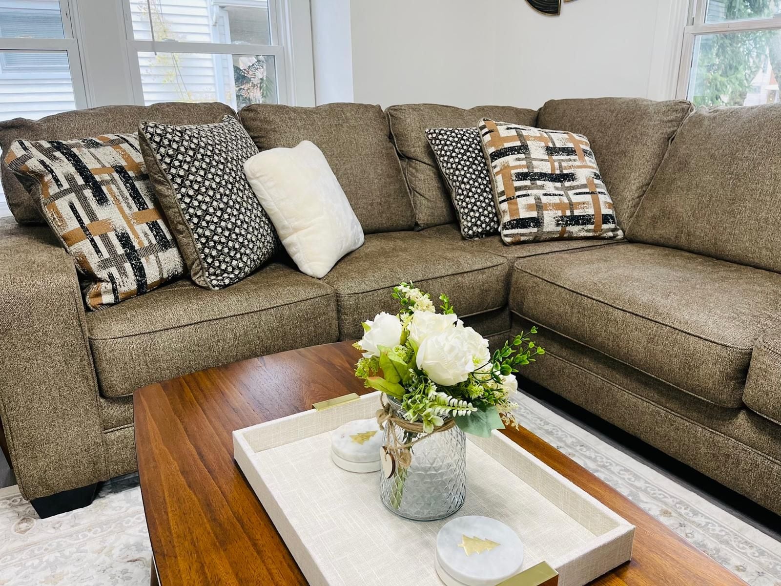 Ein Wohnzimmer mit einem braunen Ecksofa, gemusterten Zierkissen, einem Holztisch und einer Vase mit weißen Blumen.