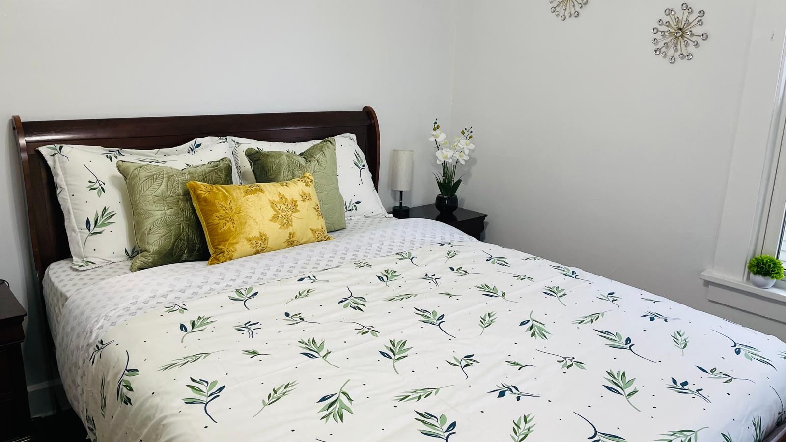 Een net opgemaakt bed met een bloemenprint deken en kussens, een nachtkastje met een lamp, en een decoratief object aan de muur.