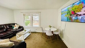 Ein Wohnzimmer mit einem dunkelbraunen Ledersofa, einem weißen Couchtisch und einem kleinen runden Tisch mit weißen Stühlen. Ein großes Fenster mit weißen Jalousien, und ein Landschaftsbild hängt an der Wand.