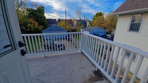 Ein Balkon mit weißer Brüstung, einem Betonboden und Blick auf angrenzende Häuser und Bäume.