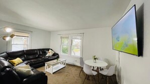 Ett vardagsrum med en svart lädersoffa, ett vitt soffbord, ett runt matbord med fyra stolar och en stor, platt tv-skärm som är monterad på väggen.