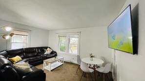 A living room with a black leather sofa, a white coffee table, a round dining table with four chairs, and a large flat-screen TV mounted on the wall. - Central West Blvd 2bedroom Apartment close to HWY (Cleveland)