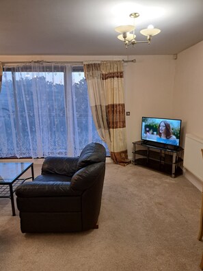 Ein Wohnzimmer mit einer Ledersofa, einem Glastisch, einem Fernseher auf einem Lowboard und einem Fenster mit Vorhängen.