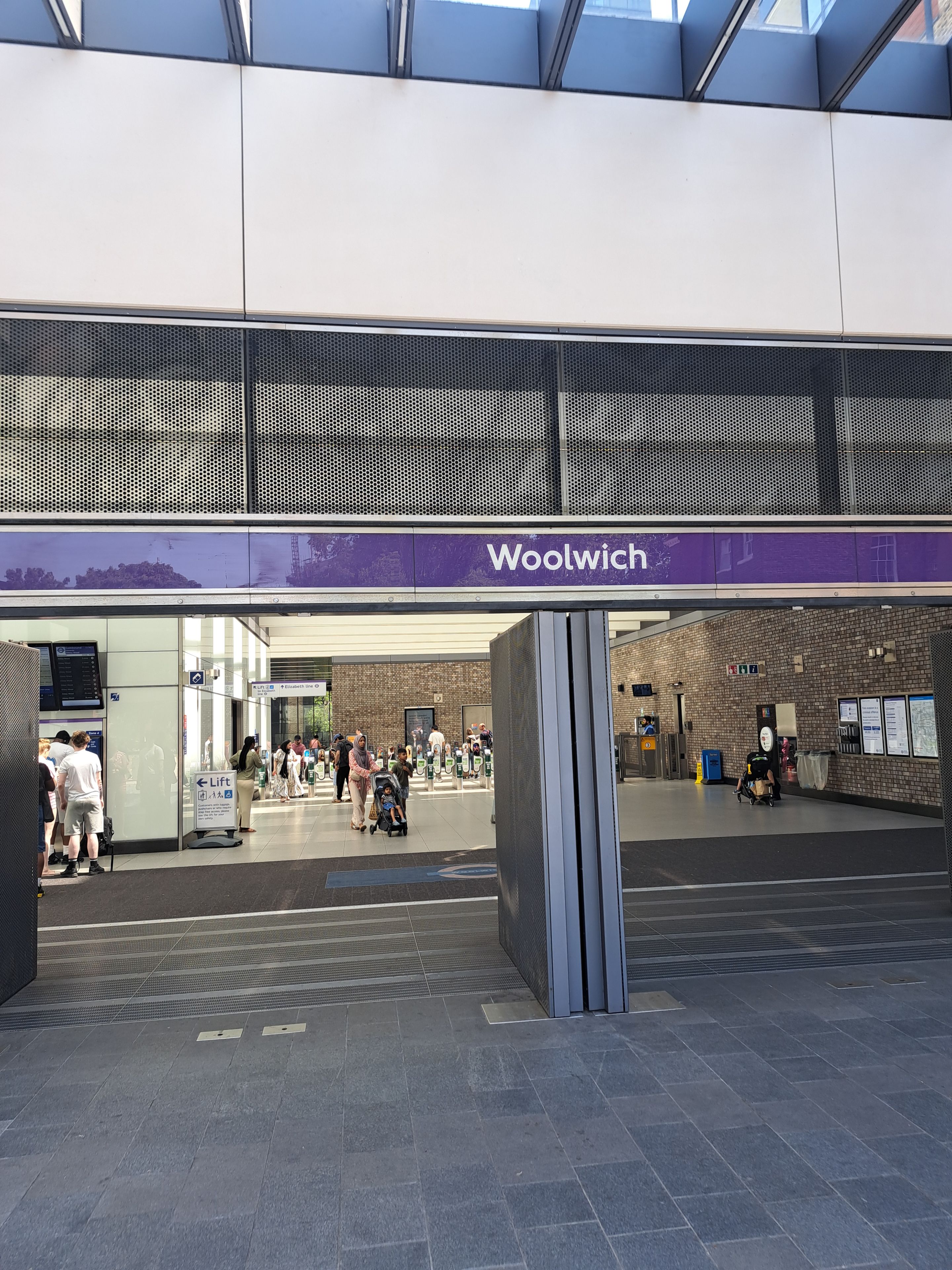 A Woolwich station entrance with a modern design, including a purple sign, a covered waiting area, and a tiled floor.