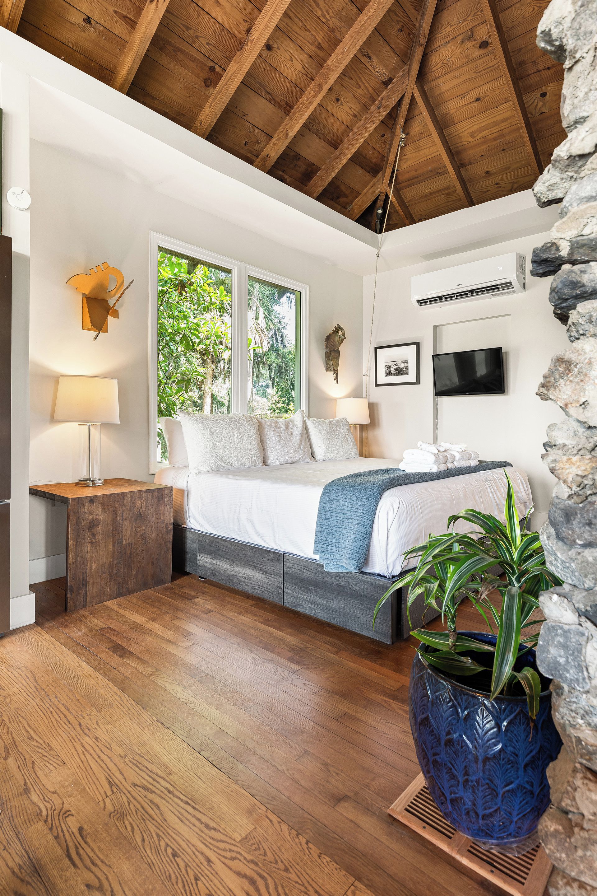 A bedroom with a wooden ceiling, a bed with white bedding, a stone wall, a TV, and a potted plant.