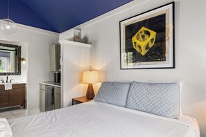 Ein Hotelzimmer mit Bett, Nachttischlampe, einem Wandbild und einem Badezimmer mit Spiegel und Waschbecken.