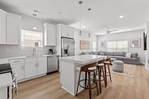 A modern kitchen with white cabinetry, a central island with bar stools, a dining area with a gray sofa, and a ceiling fan. - Electric Octopus-2.5 Blocks-Beach-Pets (Myrtle Beach)