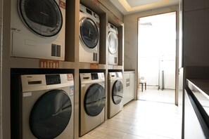 Une buanderie équipée de plusieurs machines à laver et de plusieurs sèche-linge, avec une chaise et une porte donnant sur une autre pièce.