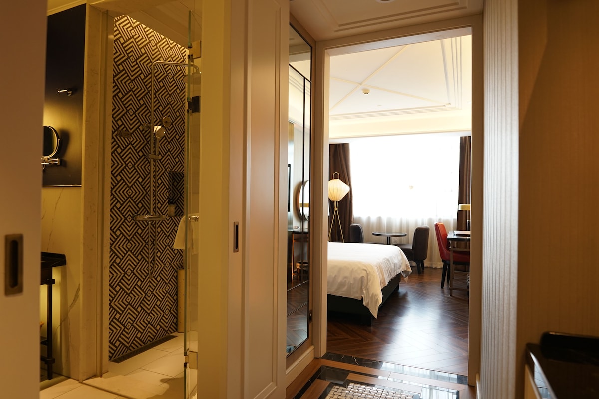 Habitación de hotel con cama, escritorio y silla. Incluye un baño con ducha y retrete. La habitación tiene un ventanal grande y una puerta que da a otra habitación.