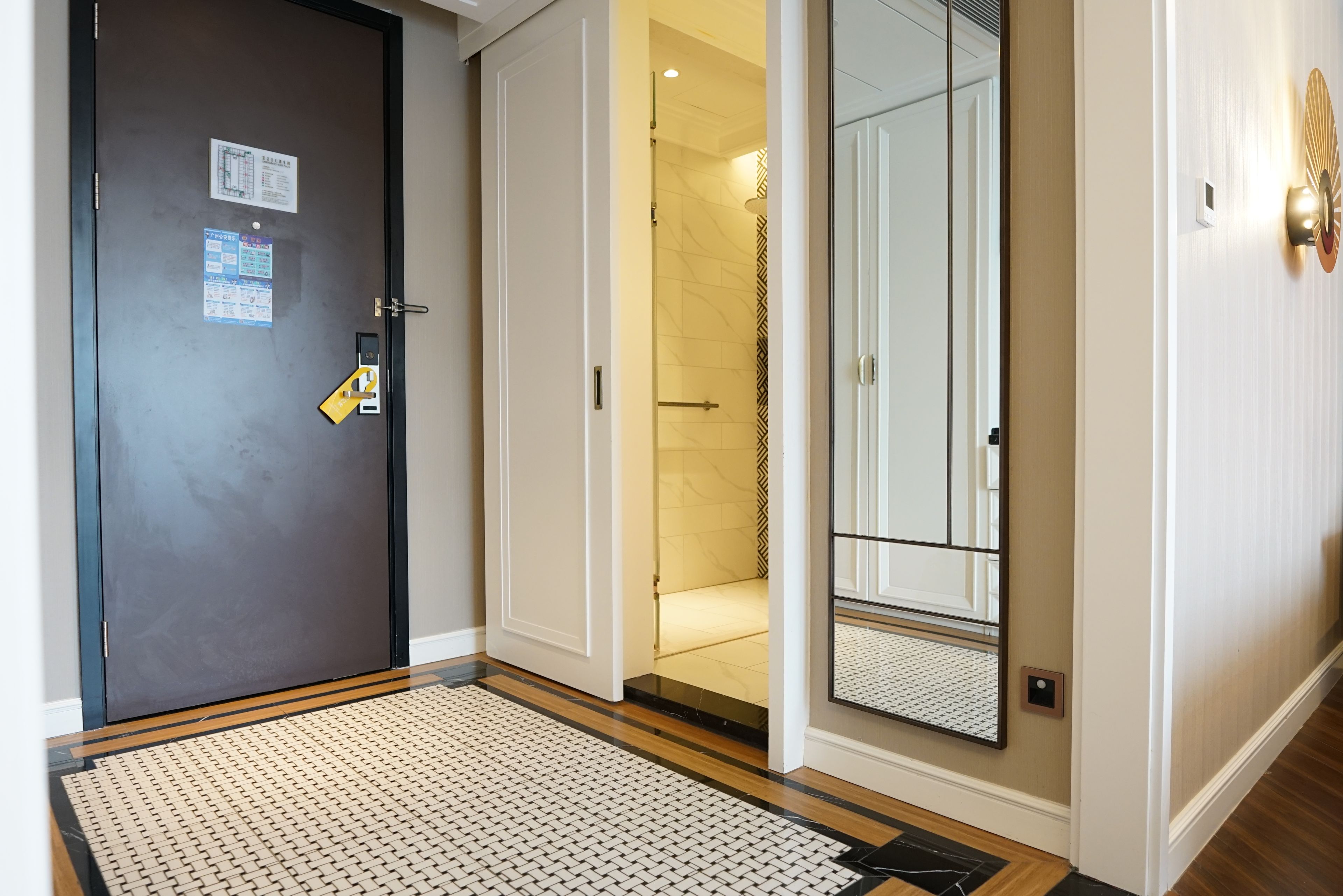 A hotel entrance with a dark door, a visible room number, a yellow keycard holder, a mirror reflecting the room, and a patterned rug.