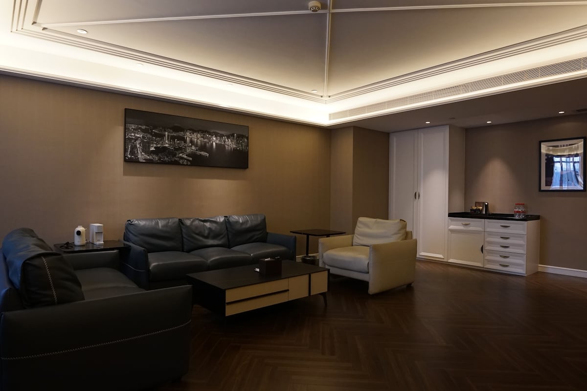 Una sala de estar moderna con sofá, sillón y una mesa de centro. Hay un cuadro en la pared y una zona de cocina al fondo.