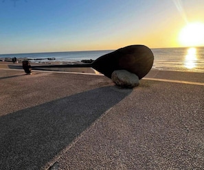 Eine Strandansicht mit einem großen, dunklen, gebogenen Objekt im Sand, einem klaren Himmel und der Sonne, die am oder über dem Horizont untergeht oder aufgeht.