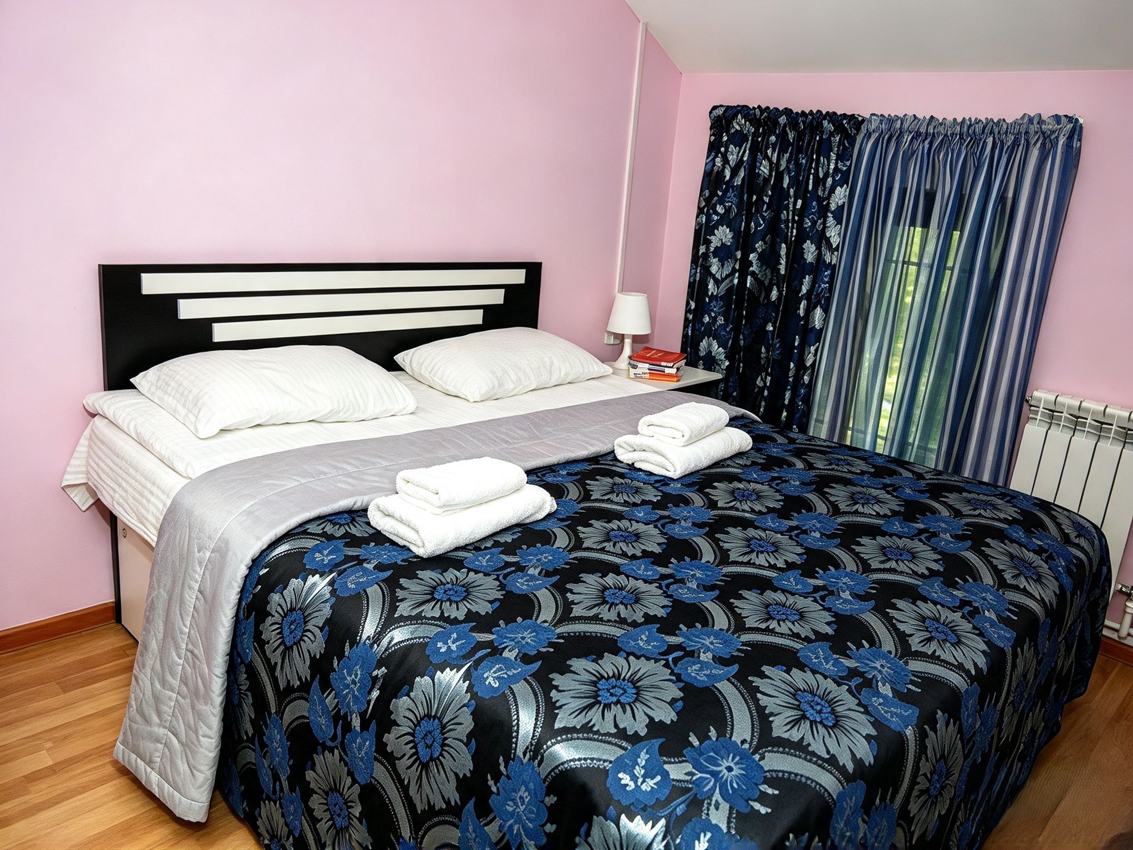 Una cama bien hecha con sábanas blancas y una colcha con estampado floral, una mesita de noche con lámpara y libros, y una ventana con cortinas azules.