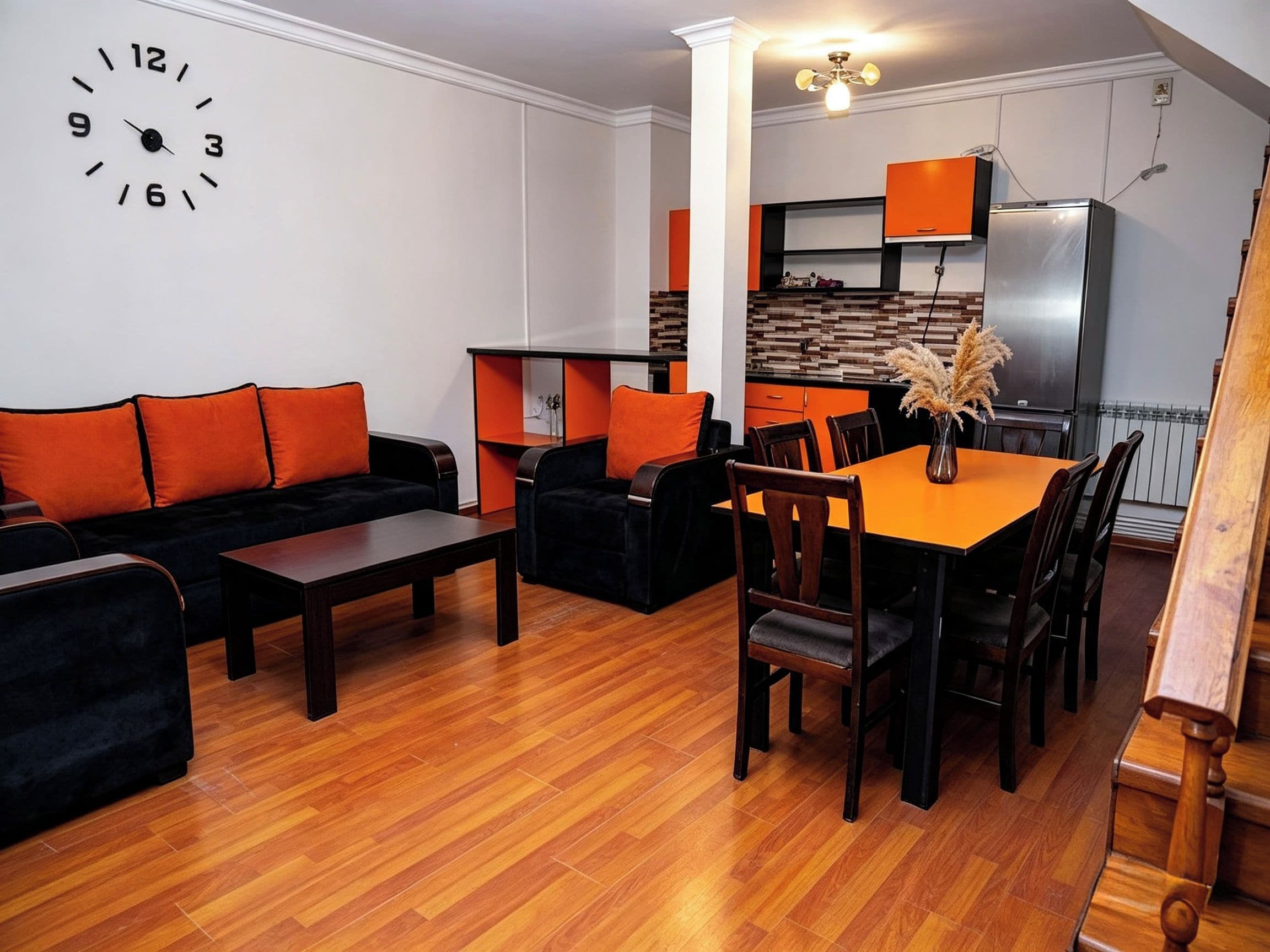 A living room with a sofa, a dining table with chairs, a kitchen area, and a clock on the wall.