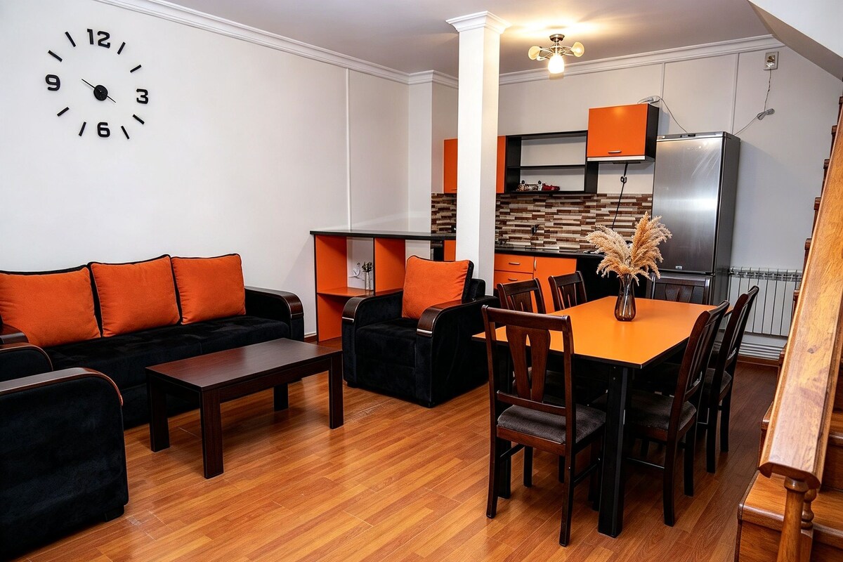 A modern living room with a black sofa, orange cushions, a wooden coffee table, and a dining area with a wooden table and chairs.