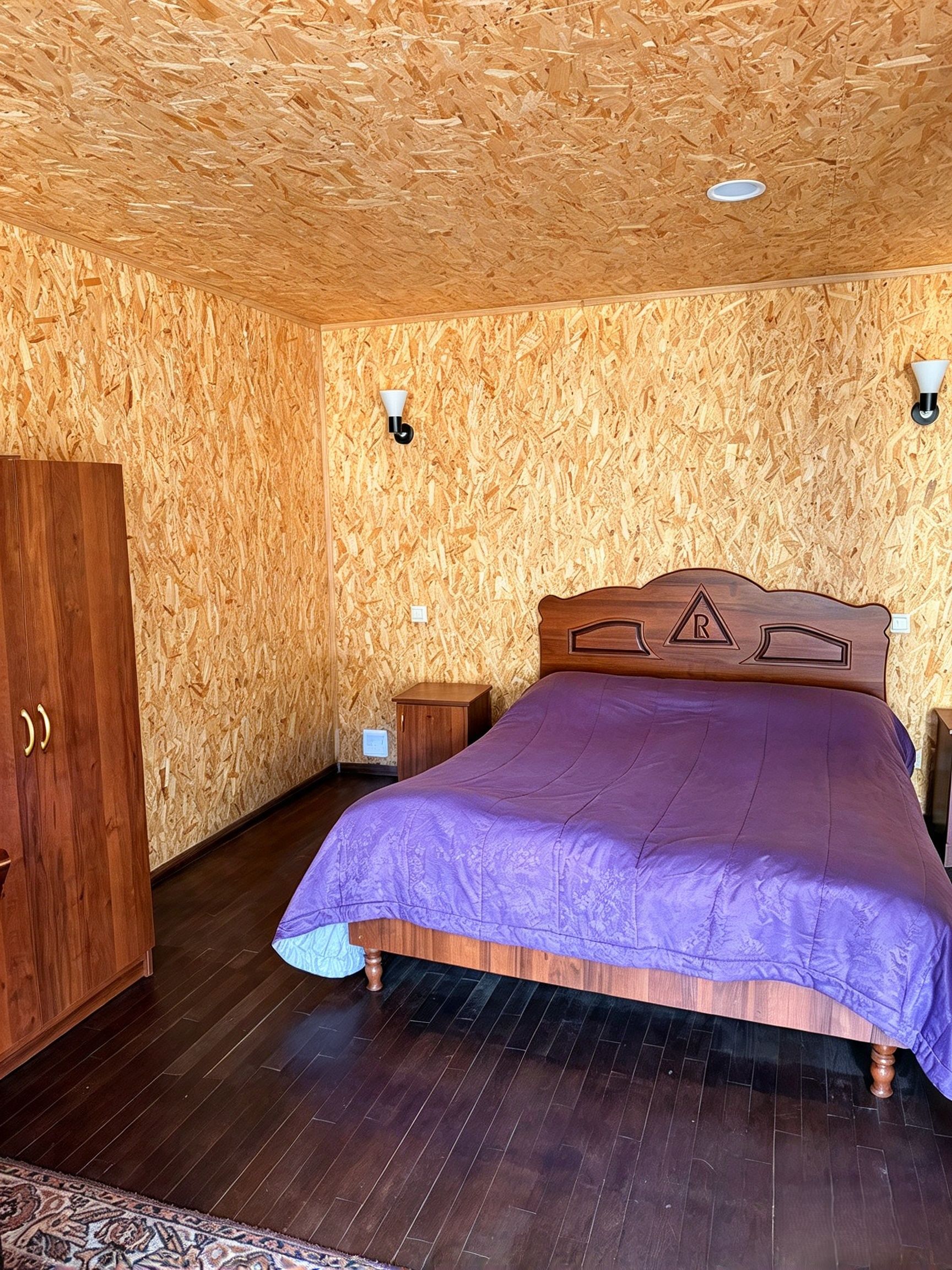 A bedroom with a wooden headboard, a bed with a purple bedspread, a wooden wardrobe, and a small bedside table.