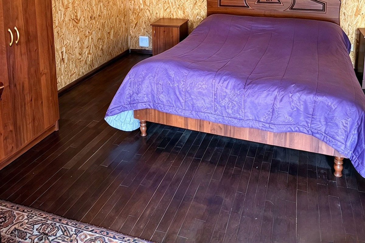 A bedroom with a wooden headboard, a bed with a purple bedspread, a wooden wardrobe, and a small bedside table.