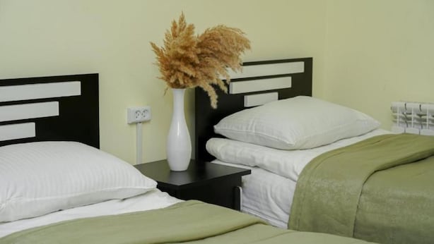 Una cama con ropa de cama blanca, un plaid verde y un florero con flores secas sobre una mesita de noche.